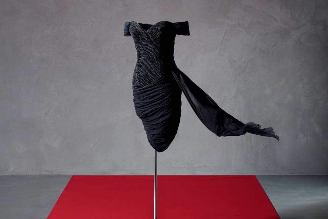 Een zwarte off-shoulder jurk met geplooide details en een lange sleep op een mannequin, geplaatst op een rode loper tegen een grijze muur.