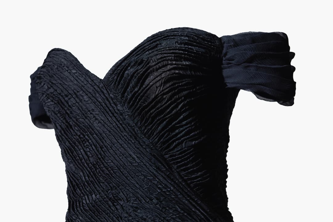 Gedetailleerde opname van een zwart kledingstuk met diagonale plooien en textiele structuur tegen witte achtergrond.