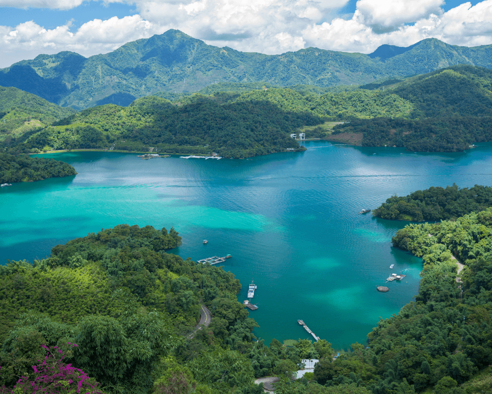 Luchtfoto van een turquoise meer omringd door dichte groene bossen, bergen en enkele boten in het water.