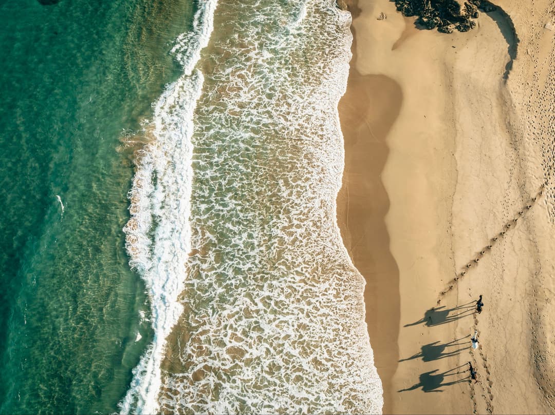 Luchtfoto van een zandstrand met drie surfers in het ondiepe water en witte schuimende golven langs de kust.